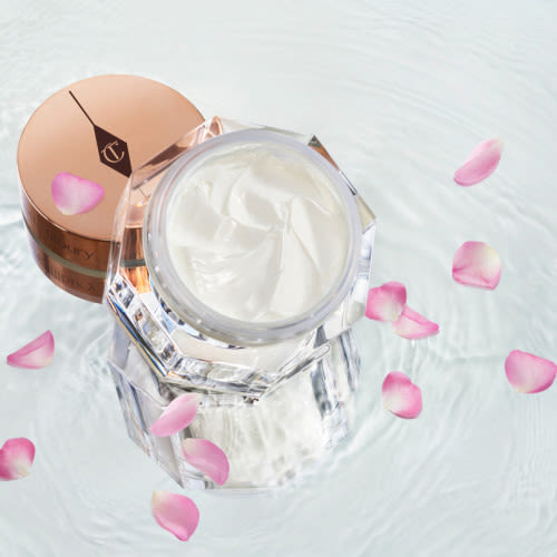 Crème épaisse de couleur blanc nacré dans un pot en verre ouvert avec son couvercle rose doré posé à côté, dans une eau cristalline où flottent des pétales de rose.