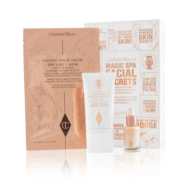 An elegant, white and gold skincare box with text in shiny gold colour that reads, 'Charlotte Tilbury's Magic Spa Facial Secrets' along with an exfoliating, wash-off mask in a white-coloured tube with a travel-size bottle of luminous face serum with a dropper lid, and a face mask in rose-gold-coloured foil packaging.