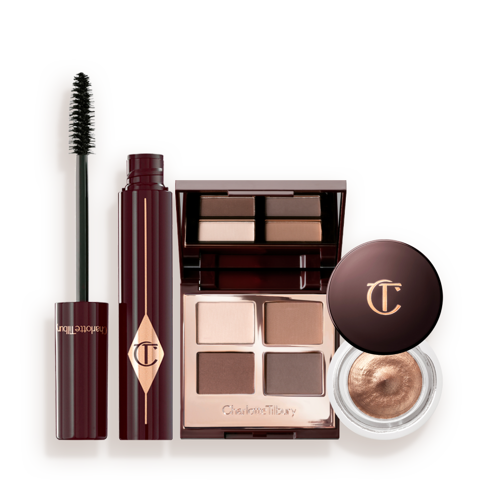 An open, mirrored-lid quad eyeshadow palette with beige, brown-gold, rose gold, and dusky pink shades, a mascara in black, and cream eyeshadow in a coppery-gold shade in a glass pot with a dark brown-coloured lid. 