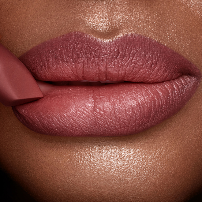 Matte Revolution Pillow Talk Medium lipstick deep tone close-up A deep tone model with glowing skin wearing a berry-pink lipstick with the lipstick gently pressed to the lower lip.
