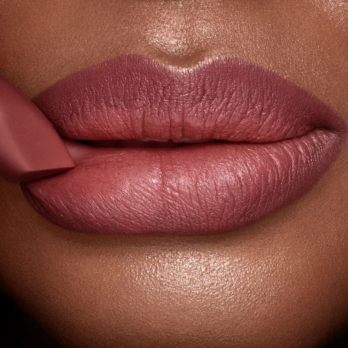 A deep tone model with glowing skin wearing a berry-pink lipstick with the lipstick gently pressed to the lower lip. 