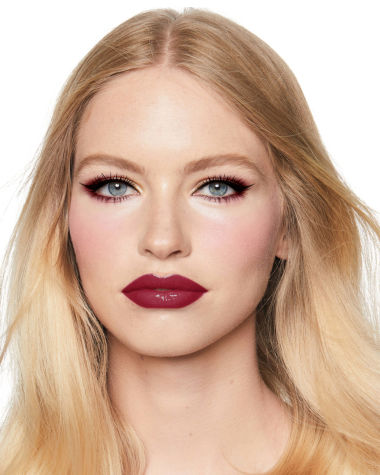 A fair-tone model with blue eyes wearing reddish-plum eye makeup, glowy face base, and a vampy-red lipstick with a satin-finish. 