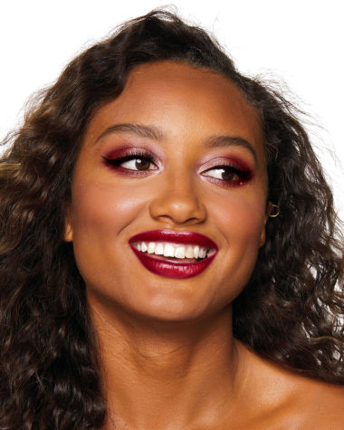 A deep-tone brunette model with reddish-plum eye makeup, glowy face base, and a vampy-red lipstick with a satin-finish. 