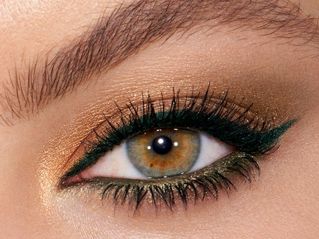 Single eye close-up of a model with hazel eyes wearing bottle-green eyeliner on her upper lid and khaki-green on her lower waterline along with shimmery gold eyeshadow. 