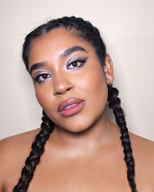 Close-up of a medium-tone model with brown eyes wearing nude pink matte lipstick with lips lined in a brown shade, bronzed skin, and ice-blue eye makeup with royal blue eyeliner. 