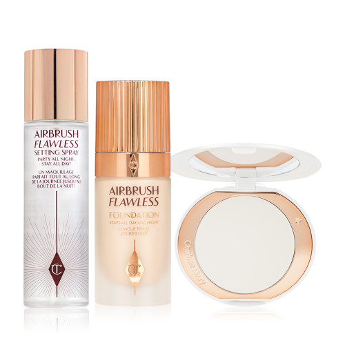 Setting spray in a large, clear bottle with a gold-coloured lid, foundation in a glass bottle with a gold-coloured lid, and pressed powder compact in white in white-coloured packaging.