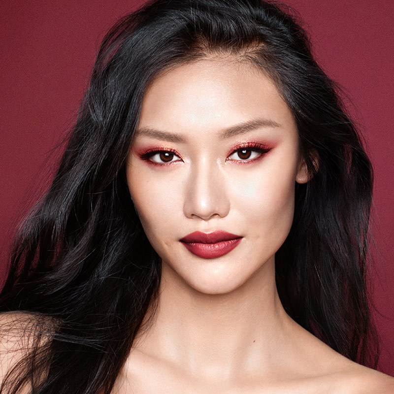 A fair-tone brunette model sporting soft red-coloured eye makeup with bold red lips and a glowy face base. 