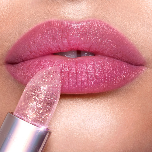 Lips close-up of a fair-tone model wearing a subtle purplish-pink glittery lipstick while holding a sparkling pink lipstick against her lips. 
