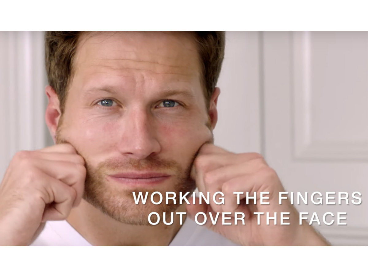 A light-tone, brunette male model massaging a face cream into the skin with knuckles.