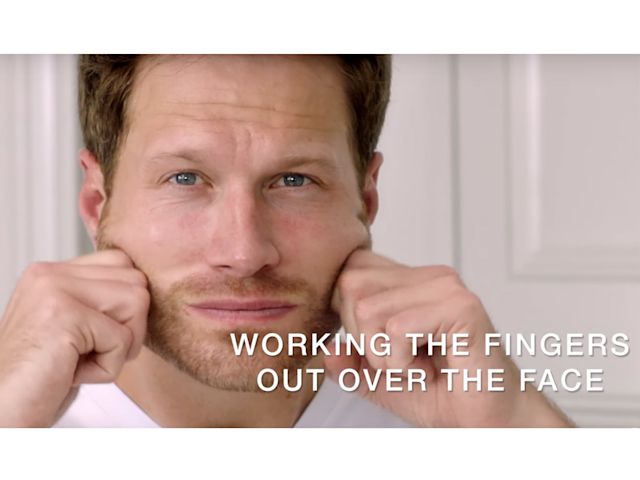 A light-tone, brunette male model massaging a face cream into the skin with knuckles.