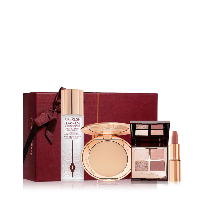 A dark scarlet-coloured gift box with a pressed powder compact with a mirrored-lid, an open lipstick in a mauve shade, a quad eyeshadow palette with pink and gold shades, and a setting spray. 