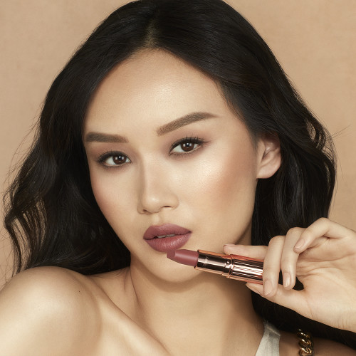 A fair-tone model wearing smokey eyeshadow with black eyeliner and a mid-toned muted nude-rose matte lipstick while holding the lipstick.
