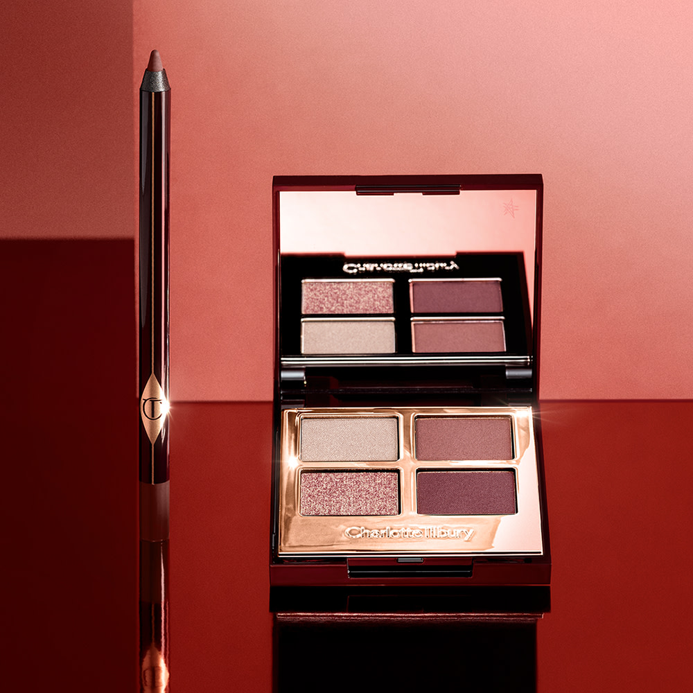 An open, mirrored-lid quad eyeshadow palette with berry-pink, red, rose gold, and beige eyeshadows and an eyeliner pencil in a dark copper shade. 
