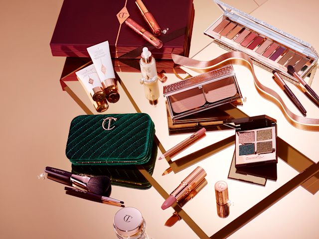 A collection of lipsticks, lip glosses, eyeshadow palettes, contour palettes, highlighter and blush palettes, primers, serums, compacts, makeup brushes, and makeup bags placed alongside a dark scarlet-coloured gift box. 