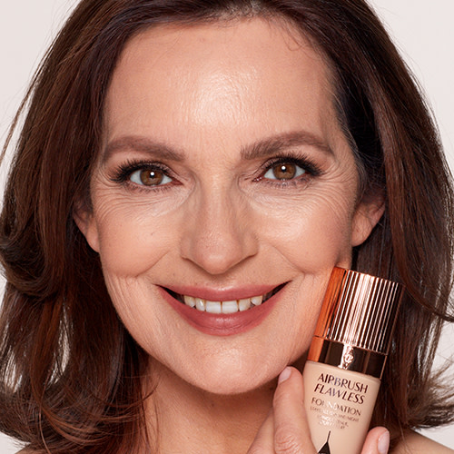 A light-tone brunette model with mature skin wearing natural-looking, glowy makeup with a flawless face base and holding a foundation bottle to her face. 