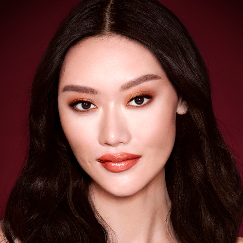 A fair-tone model with brown eyes wearing shimmery copper and gold eye makeup with black eyeliner, glowy bronzed cheeks, and orange-red lipstick with gloss on top. 