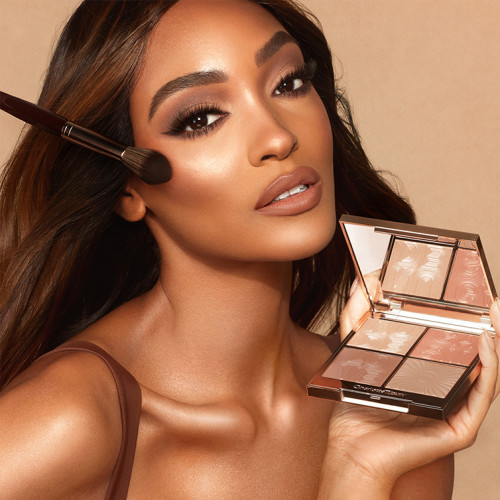 Deep-tone brunette model with brown eyes and glowy, bronzed skin, applying peach highlighter blush from a quad face palette with contouring, eyeshadow, and highlighter in nude shades.