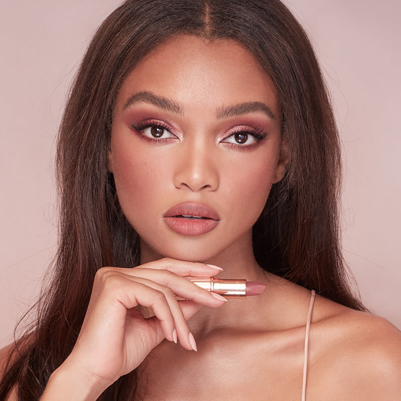 Deep-tone model with brown eyes wearing nude pink matte lipstick with warm berry-pink blush, shimmery rose gold eye makeup, and candlelight-gold highlighter subtly applied on the high points of her face. 