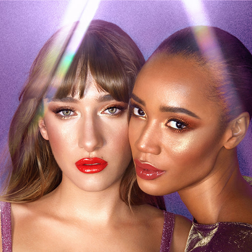Two models, one deep-tone and the other fair, wearing shimmery gold eye makeup with bold red lips. 
