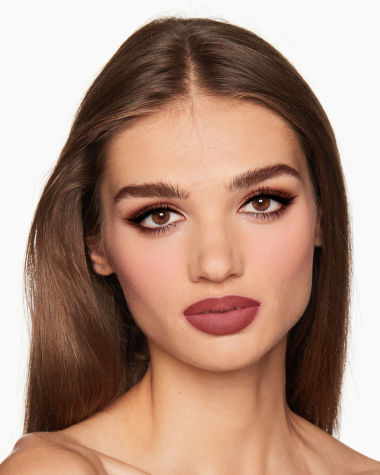 A medium-tone model with brown eyes wearing shimmery brown and gold eyeshadow with black kohl liner on her upper lid and lower waterline, a cool-toned glowy pink blush, and a berry-pink matte lipstick. 