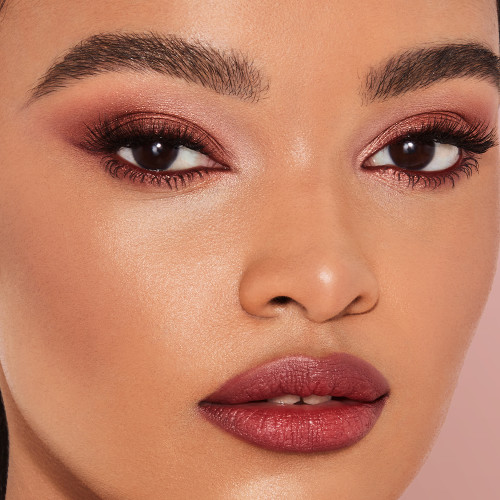 Close-up of a deep-tone model with brown eyes wearing a shimmery russet rose cream eyeshadow with a golden sparkle, dark brown eyeliner, and lipstick in a berry-pink shade with matching lip liner. 