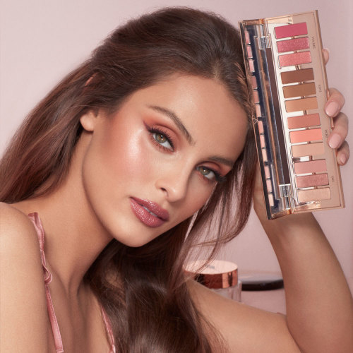 A light skin, brunette model wearing glowy peach and pink eye makeup with berry-pink lips and terracotta blush while holding an eyeshadow palette with nude pink, peach, and brown shades.