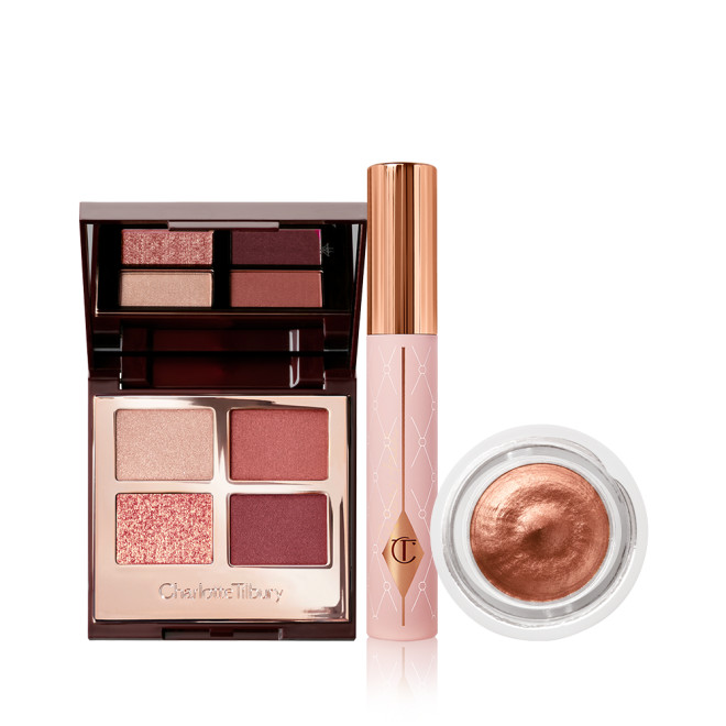 An open, quad eyeshadow palette with a mirrored-lid in red, pink, and gold shades, mascara in a nude pink tube with a gold-coloured lid, and cream eyeshadow in a glass pot in a coppery-gold shade.
