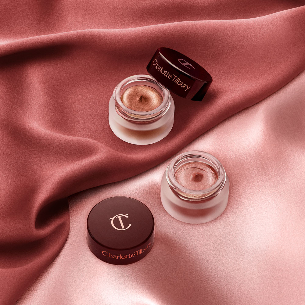 Two cream eyeshadows in glass pots, one a russet rose colour with a golden-peach sparkle and the other a nude pink with fine shimmer, with their lids placed next to them.