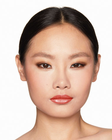 A fair-tone model with brown eyes wearing smokey brown eye makeup with warm bronze and pink blush, and glossy terracotta lips.