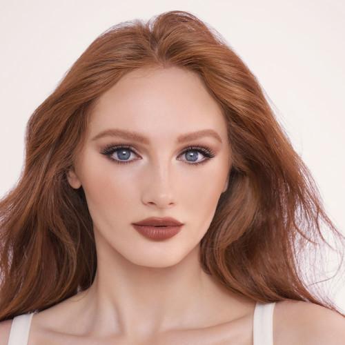Fair-tone model with blue eyes wearing soft beige and champagne eyeshadow with black eyeliner on her eyelid and nude beige eyeliner on her lower waterline along with a deep brick-red lipstick.