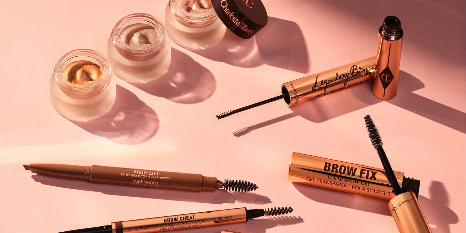 Trio of cream eyeshadows in shimmery shades of gold and pink in open pots with dark brown lids, eyebrow tints, eyebrow gels and brushes, mascara, and makeup remover in a large, clear bottle with a white-coloured cap.