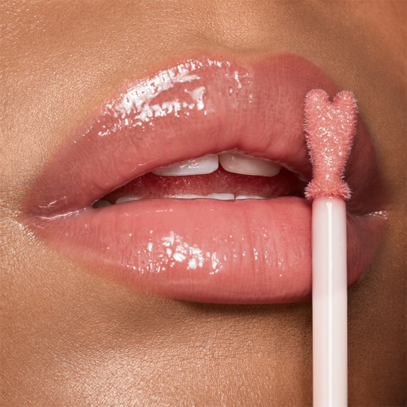 Lips close-up of a light-tone model applying sheer, nude pink lip gloss with a heart-shaped applicator. 