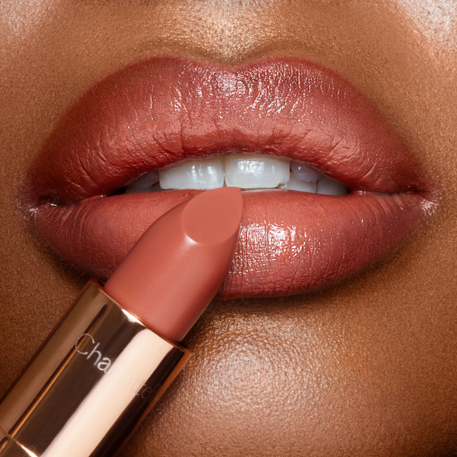 Lips close-up of a deep-tone model wearing a warm coral lipstick with a satin-finish while holding the lipstick in front of her lips.