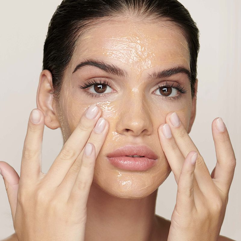 A light-tone model massaging a yellow-tinted oil cleanser on her face. 