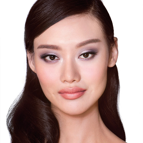 A fair-tone model with brown eyes wearing shimmery lilac and champagne eye makeup with nude pink blush and coral lipstick with gloss on top.