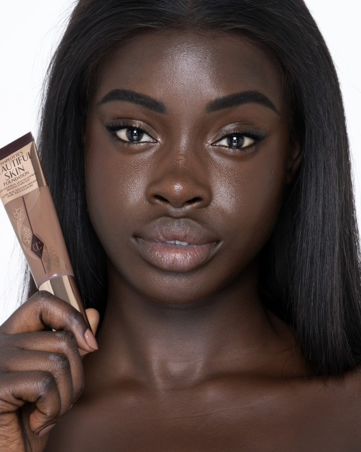 Deep-tone brunette model with brown eyes wearing nude pink lipstick with a skin-like foundation with a flawless, dewy finish. 