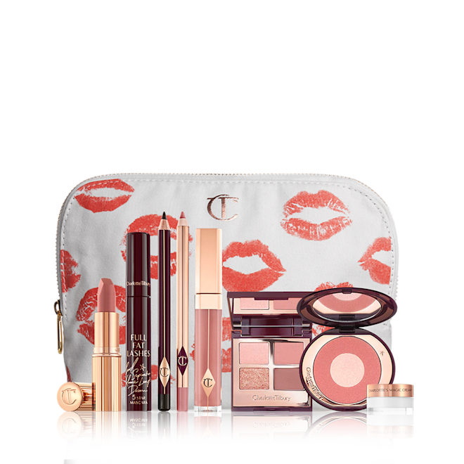 A mascara with an open, mirrored-lid quad eyeshadow palette in shimmery neutral shades, a shimmery nude pink lipstick, a nude pink lip gloss and lip liner pencil, and a two-tone powder blush compact in a bright pink shade, and a mini face cream in a glass jar with all products in front of a white-coloured makeup pouch. 