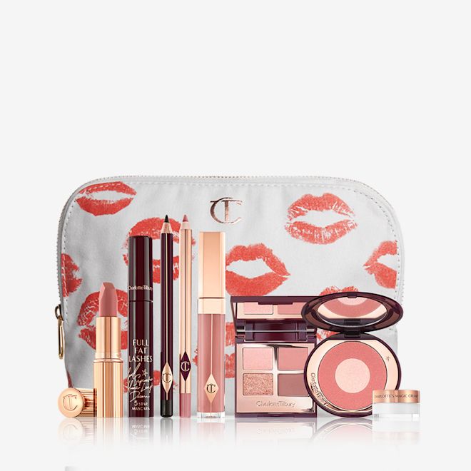 The Pillow Talk Look with mini Magic Cream displayed A mascara with an open, mirrored-lid quad eyeshadow palette in shimmery neutral shades, a shimmery nude pink lipstick, a nude pink lip gloss and lip liner pencil, and a two-tone powder blush compact in a bright pink shade, and a mini face cream in a glass jar with all products in front of a white-coloured makeup pouch.