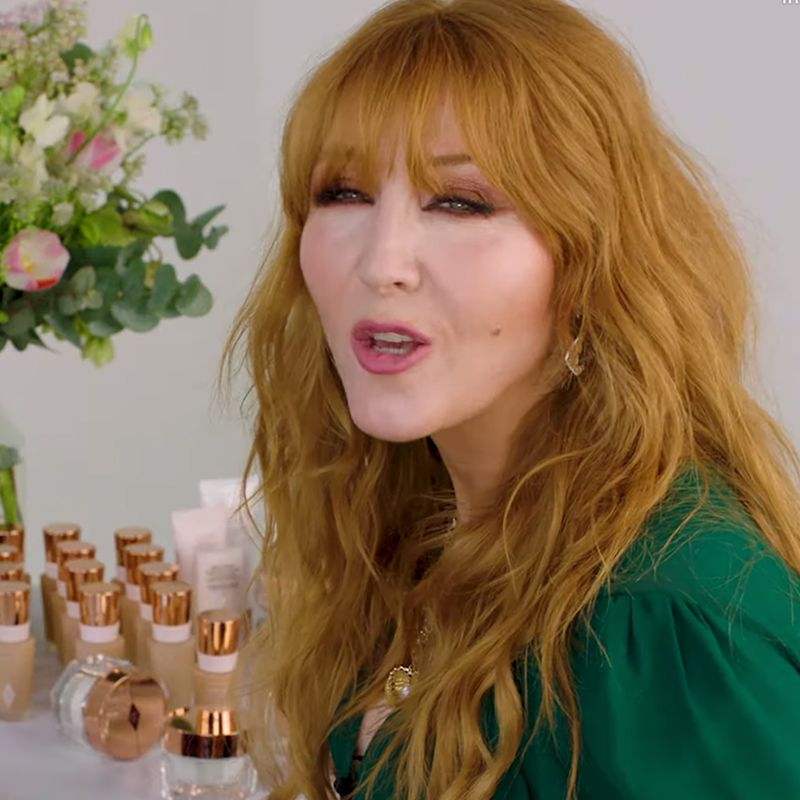 Charlotte Tilbury wearing shimmery brown eyeshadow with berry-pink lipstick while standing in front of a table covered with makeup. 