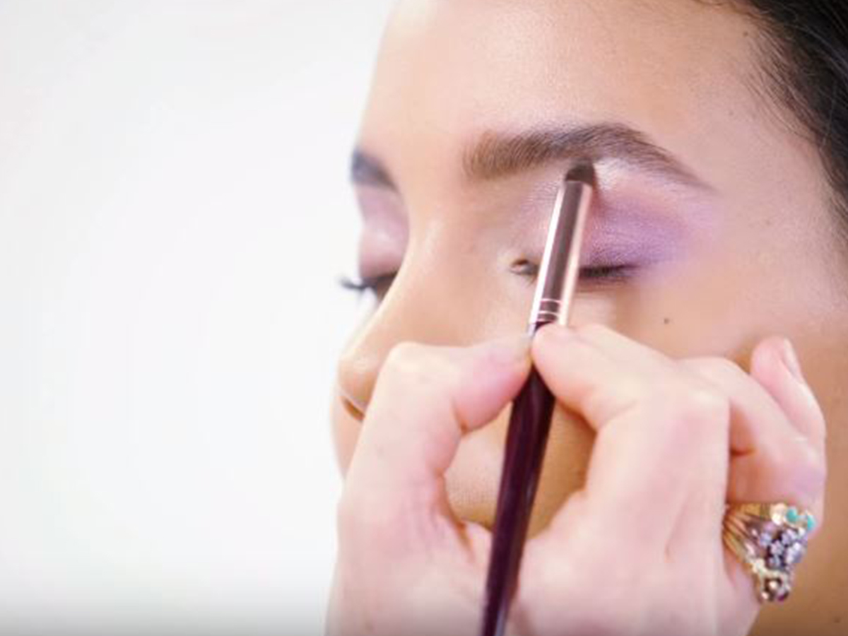 A light-tone brunette model with dewy skin wearing frosted lilac eye makeup and getting her brow bone highlighted with a shimmery cream shade. 