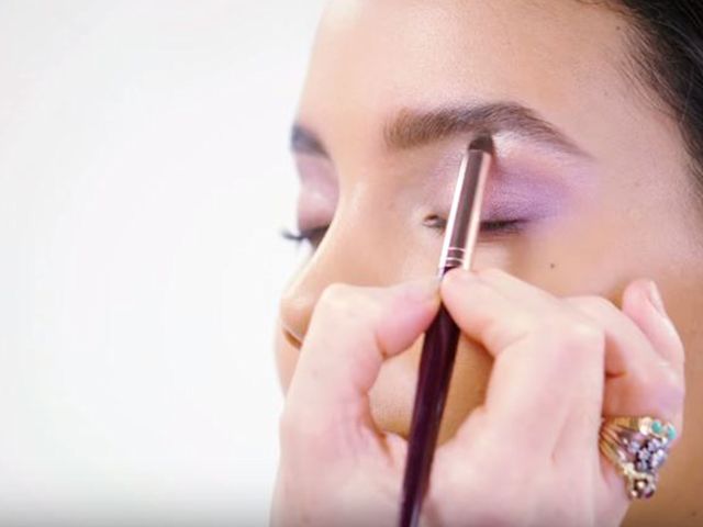 A light-tone brunette model with dewy skin wearing frosted lilac eye makeup and getting her brow bone highlighted with a shimmery cream shade. 
