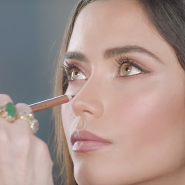 A fair-tone model with green eyes getting freckles drawn on with a light brown eyeliner pencil. 