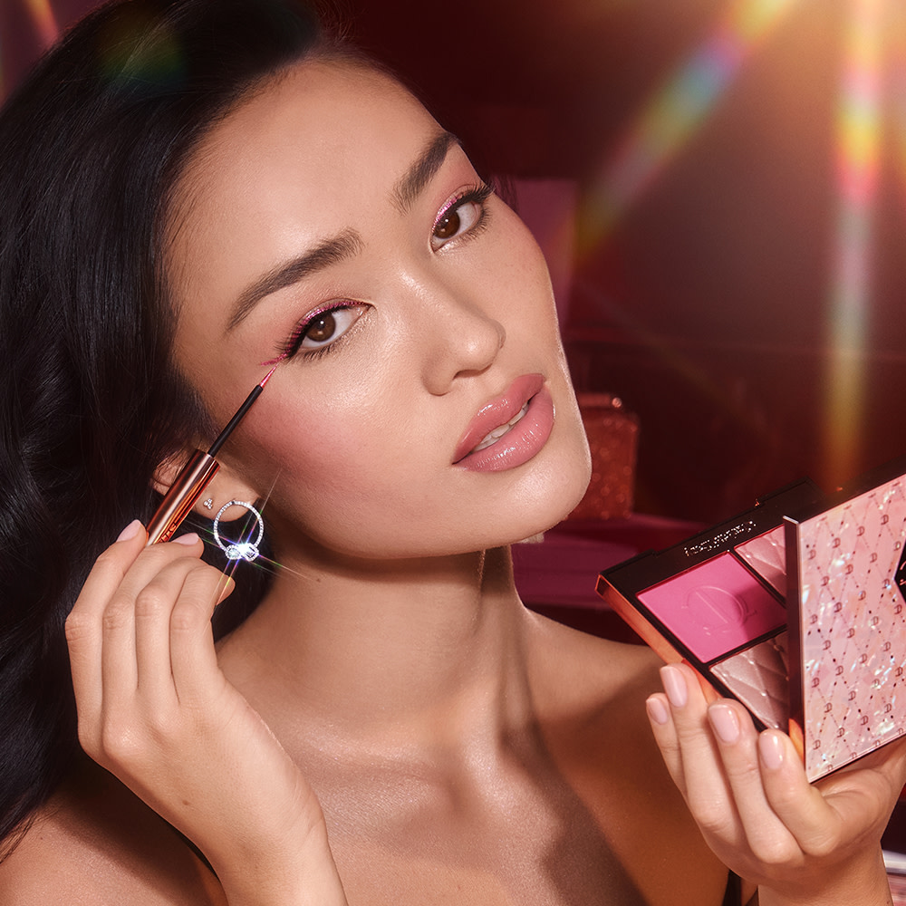 Fair-tone brunette model with brown eyes wearing full-glam, extremely glowy pink makeup while holding an open, face palette with matte and shimmery eyeshadows, blushes and highlighters in shades of pink and gold with a mirrored lid.
