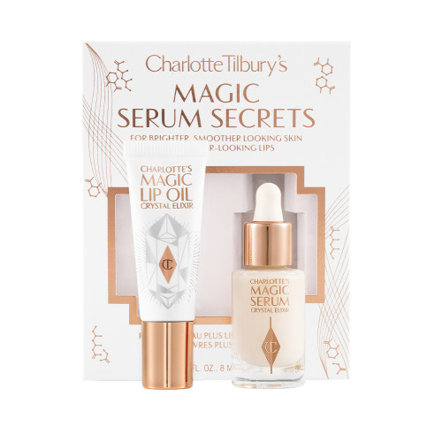 Travel-size lip oil in a white-coloured tube with a gold-coloured lid and a mini, glow-enhancing serum in a glass bottle with a gold and white-coloured dropper lid with their packaging sleeve behind them in a white colour with the text, 'Magic serum secrets' written on it'