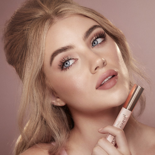 A fair-tone model with blue eyes wearing nude pink lipstick with muted pink blush, and lengthening mascara in a berry-brown shade. 