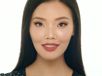 Close-up of a light-tone, brunette model with a glowing face, wearing a muted red lipstick, and black eyeliner. 