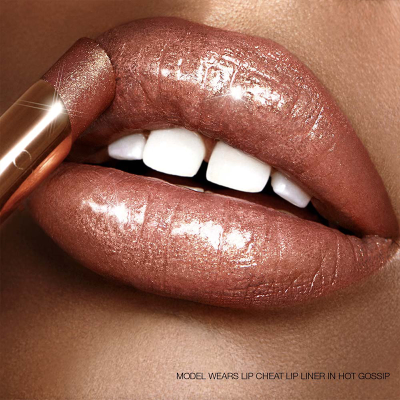 Lips close-up of a deep-tone model applying a sheer, glittery, bronze-pink lipstick. 