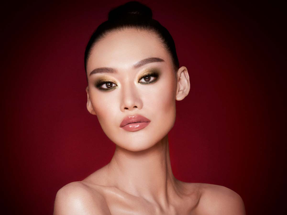 A fair-tone model with brown eyes wearing smokey green eye makeup with black eyeliner, soft brown blush, and nude pink lipstick topped up with gloss. 