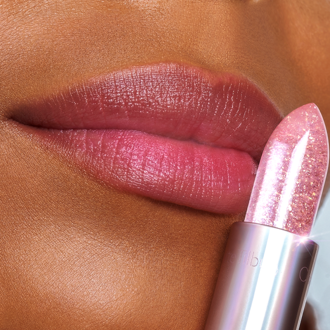 Lips close-up of a deep-tone model wearing a subtle purplish-pink glittery lipstick while holding a sparkling pink lipstick against her lips. 