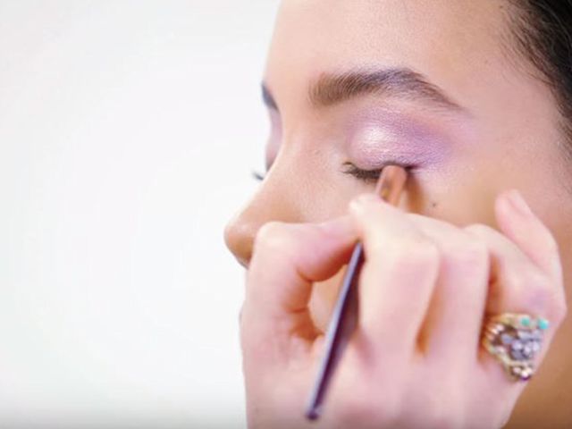 A light-tone model wearing frosted lilac eye makeup and getting smokey eyeliner applied. 